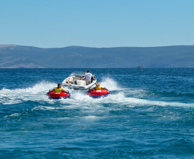 Ein Motorboot fährt über das klare blaue Wasser, während zwei Personen auf roten Bananenbooten dahinter gezogen werden. Im Hintergrund sind sanfte Hügel und ein strahlend blauer Himmel zu sehen, was eine entspannte Urlaubsatmosphäre vermittelt.