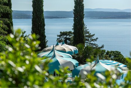 Blick auf eine ruhige Küstenlandschaft mit blauen und weißen Sonnenschirmen, umgeben von üppigem Grün und hohen Zypressen. Im Hintergrund erstreckt sich das glitzernde Wasser des Sees unter einem k... Blick auf eine ruhige Küstenlandschaft mit blauen und weißen Sonnenschirmen, umgeben von üppigem Grün und hohen Zypressen. Im Hintergrund erstreckt sich das glitzernde Wasser des Sees unter einem klaren Himmel.