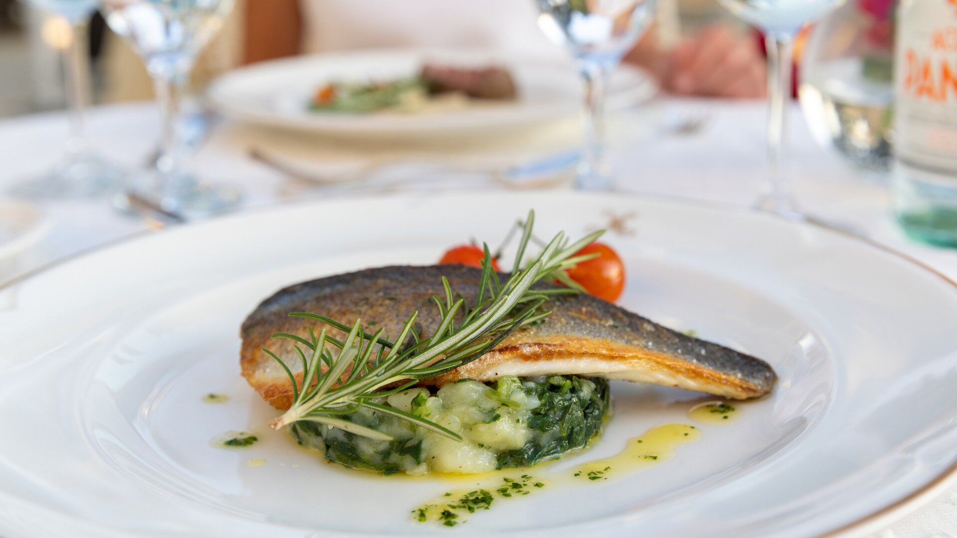 Ein kunstvoll angerichteter Teller mit gebratenem Fisch, garniert mit frischem Rosmarin und Kirschtomaten. Darunter liegt ein Bett aus cremigem Kartoffelpüree und Spinat. Im Hintergrund sind weitere Teller und Gläser sichtbar, die eine elegante Atmosphäre schaffen.