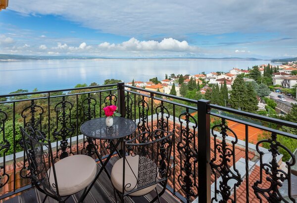Balkon mit Blick auf eine ruhige Bucht, umgeben von grünen Bäumen und roten Dächern. Ein kleiner Tisch mit zwei Stühlen und einer Vase mit Blumen lädt zum Entspannen ein. Der Himmel ist teilweise b... Familienurlaub Kroatien ist so schön im Kvarner Palace