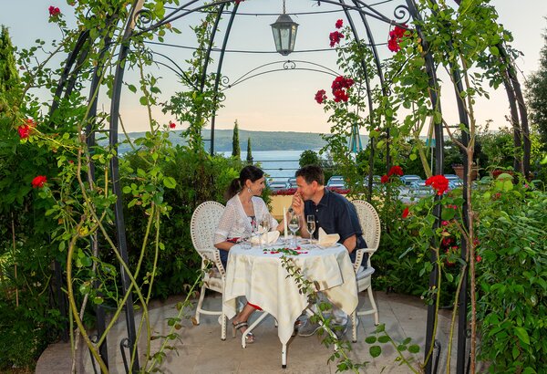 Ein romantisches Dinner für zwei unter einem Rosenbogen, umgeben von üppigem Grün. Der Tisch ist elegant gedeckt, und das Paar genießt Wein mit Blick auf das Wasser im Hintergrund. Die Atmosphäre ist entspannt und einladend.