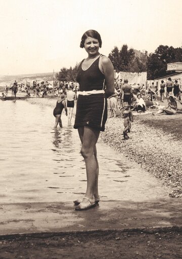 Ein Mädchen in einem Badeanzug steht am Ufer eines Sees. Im Hintergrund sind viele Menschen beim Schwimmen und Entspannen am Strand zu sehen. Die Szene vermittelt eine nostalgische Sommeratmosphäre.