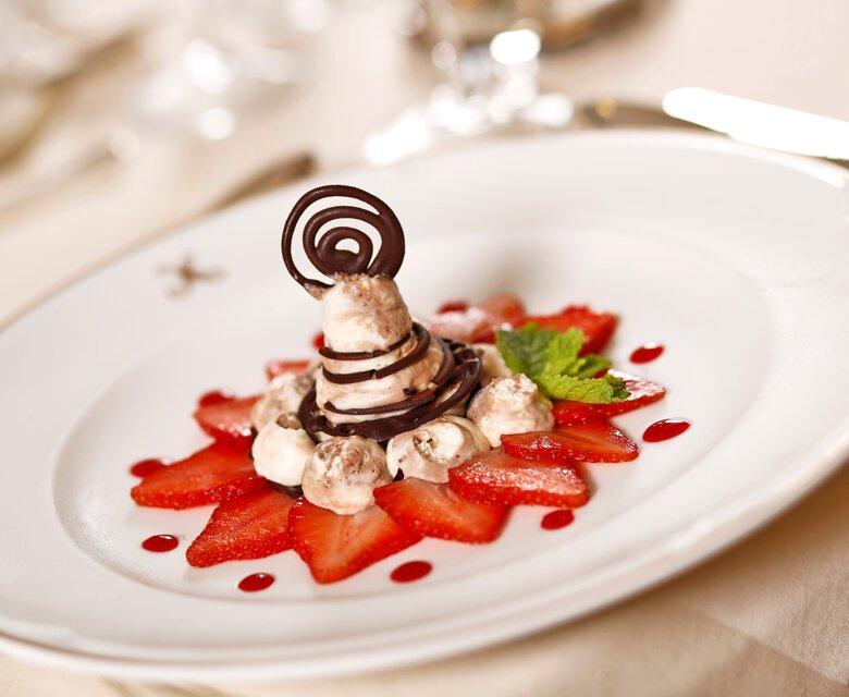 Ein kunstvoll angerichtetes Dessert auf einem weißen Teller, bestehend aus einer Schokoladenspirale, Sahne und frischen Erdbeeren. Rote Fruchtsauce umrahmt die Erdbeeren, und ein Blatt Minze verleiht dem Gericht eine frische Note.