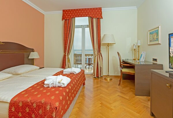 Ein helles Hotelzimmer mit zwei Einzelbetten, einem roten Bettüberwurf und frischen Handtüchern. Ein Schreibtisch und ein Fernseher sind vorhanden. Große Fenster mit Vorhängen bieten einen Blick na... Doppelzimmer mit Meerblick im Strandhotel Crikvenica