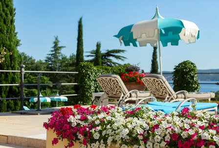 Ein sonniger Poolbereich mit bequemen Liegen und einem bunten Sonnenschirm. Im Vordergrund blühen leuchtende Petunien in Rot und Weiß. Im Hintergrund sind grüne Bäume und eine ruhige Wasserfläche s... Ein sonniger Poolbereich mit bequemen Liegen und einem bunten Sonnenschirm. Im Vordergrund blühen leuchtende Petunien in Rot und Weiß. Im Hintergrund sind grüne Bäume und eine ruhige Wasserfläche sichtbar, die eine entspannte Atmosphäre schaffen.