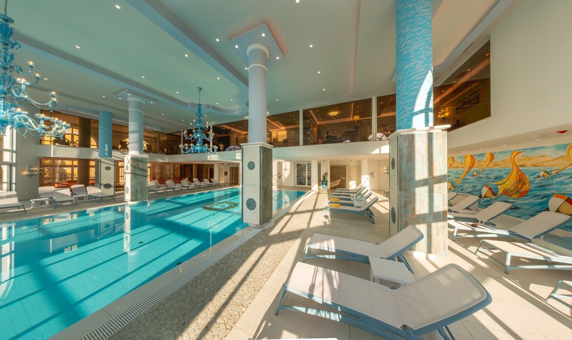 Innenansicht eines eleganten Schwimmbereichs mit einem großen, blauen Pool. Umgeben von Liegen und hellen, modernen Möbeln. Große Fenster lassen viel Tageslicht herein, während dekorative Kronleuchter und eine Wandmalerei mit maritimen Motiven für eine entspannte Atmosphäre sorgen.