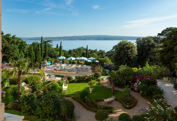 Blick auf einen gepflegten Garten mit bunten Blumen und Palmen, umgeben von Bäumen. Im Hintergrund ist ein Poolbereich mit Liegen und Sonnenschirmen zu sehen, sowie das glitzernde Wasser des Meeres... Hotel mit Meerblick in der Kvarner Bucht - Doppelzimmer