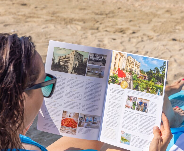 Eine Frau in einem blauen Bikini sitzt am Strand und liest einen Reiseführer. Im Hintergrund ist das Meer zu sehen, während sie auf einem gestreiften Handtuch liegt.