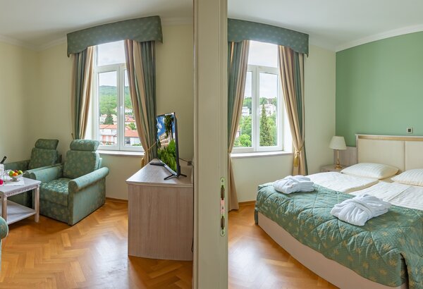 Ein helles Hotelzimmer mit zwei Bereichen: links ein Sitzbereich mit zwei bequemen Stühlen und einem Tisch, rechts ein Schlafzimmer mit einem Doppelbett und einem Nachttisch. Große Fenster bieten e... Familienurlaub Kroatien ist so schön im Kvarner Palace