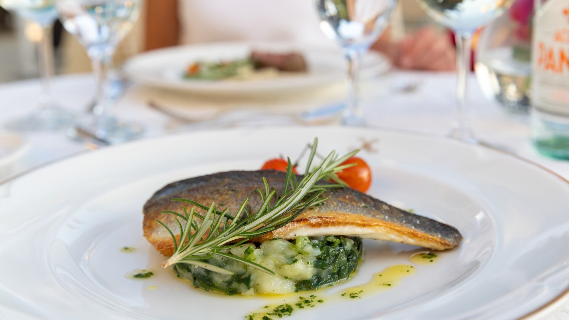Ein kunstvoll angerichteter Teller mit gebratenem Fisch, garniert mit frischem Rosmarin und Kirschtomaten, serviert auf einem Bett aus püriertem Gemüse. Im Hintergrund sind Gläser mit Wasser und ein weiterer Teller sichtbar, was eine elegante Essensatmosphäre schafft.