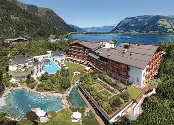 Ein malerisches Hotel mit mehreren Gebäuden, umgeben von üppigen Gärten und einem klaren Pool. Im Hintergrund erstreckt sich ein schöner See, umgeben von Bergen. Die Atmosphäre ist ruhig und einladend, ideal für einen entspannten Urlaub.
