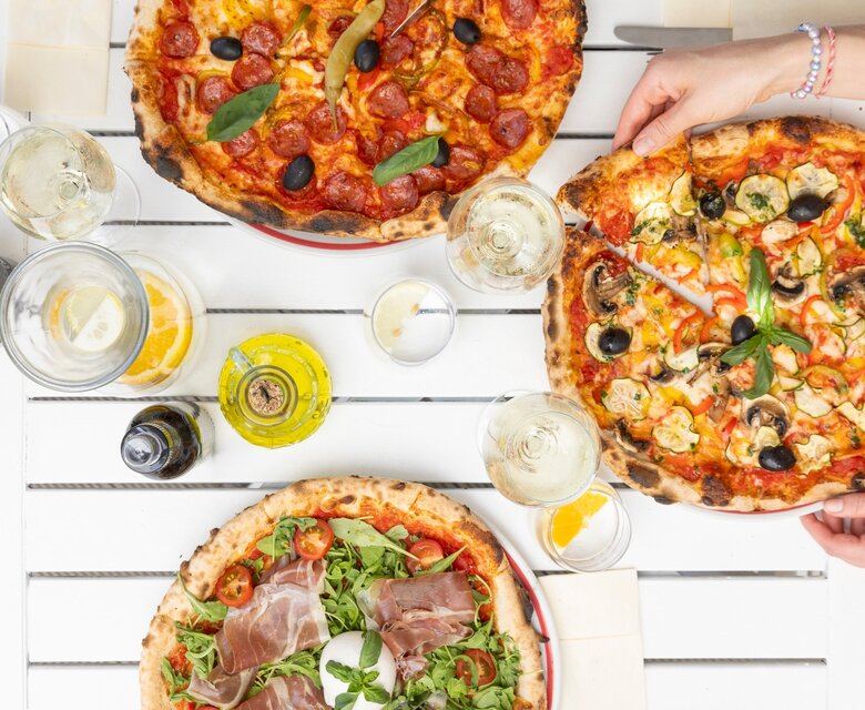 Drei köstliche Pizzen auf einem weißen Tisch, umgeben von Gläsern mit Getränken. Hände greifen nach den Pizzen, die mit verschiedenen Belägen wie Schinken, Gemüse und Oliven belegt sind. Eine einladende, gesellige Atmosphäre.