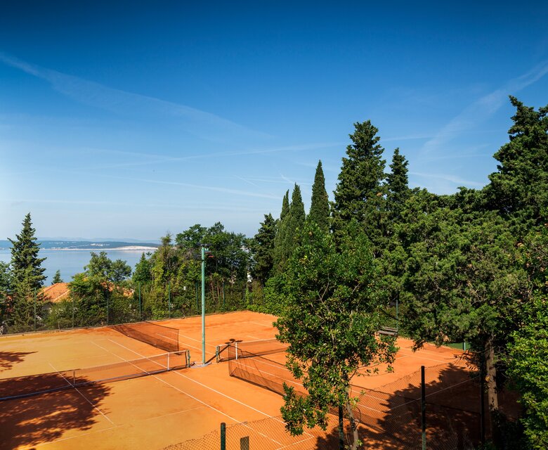 Tennisplatz mit rotem Sand, umgeben von üppigem Grün und hohen Bäumen. Im Hintergrund erstreckt sich ein Blick auf das Wasser und den klaren blauen Himmel.