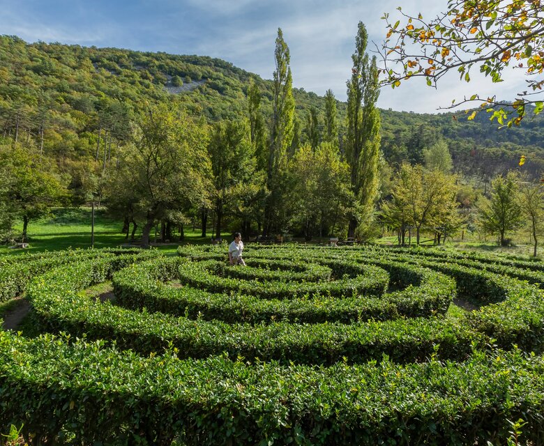 Ein weitläufiger Garten mit einem spiralförmigen Heckenlabyrinth, umgeben von üppigem Grün und hohen Bäumen. Im Hintergrund sind sanfte Hügel zu sehen, während eine Person im Labyrinth steht und die ruhige, natürliche Atmosphäre genießt.