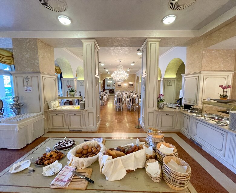 Ein eleganter Speisesaal mit einem Buffetbereich im Vordergrund, der mit verschiedenen Brotsorten und Gebäck gefüllt ist. Im Hintergrund sind Tische mit Stühlen und ein Kronleuchter sichtbar, was eine einladende Atmosphäre schafft.