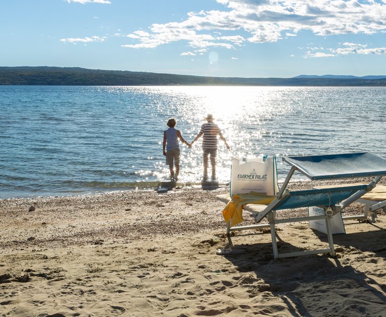 Ein Paar hält Händchen und steht am Ufer eines ruhigen Strandes, während die Sonne auf das Wasser scheint. Im Vordergrund sind Liegen auf dem Sand zu sehen, und im Hintergrund erstreckt sich eine sanfte Küstenlandschaft.