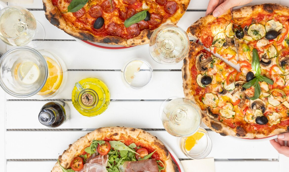 Drei verschiedene Pizzen auf einem weißen Tisch, umgeben von Gläsern mit Getränken. Eine Pizza mit Salami und Basilikum, eine mit Meeresfrüchten und Gemüse, und eine mit Rucola und Schinken. Einladende Atmosphäre mit frischen Zutaten.