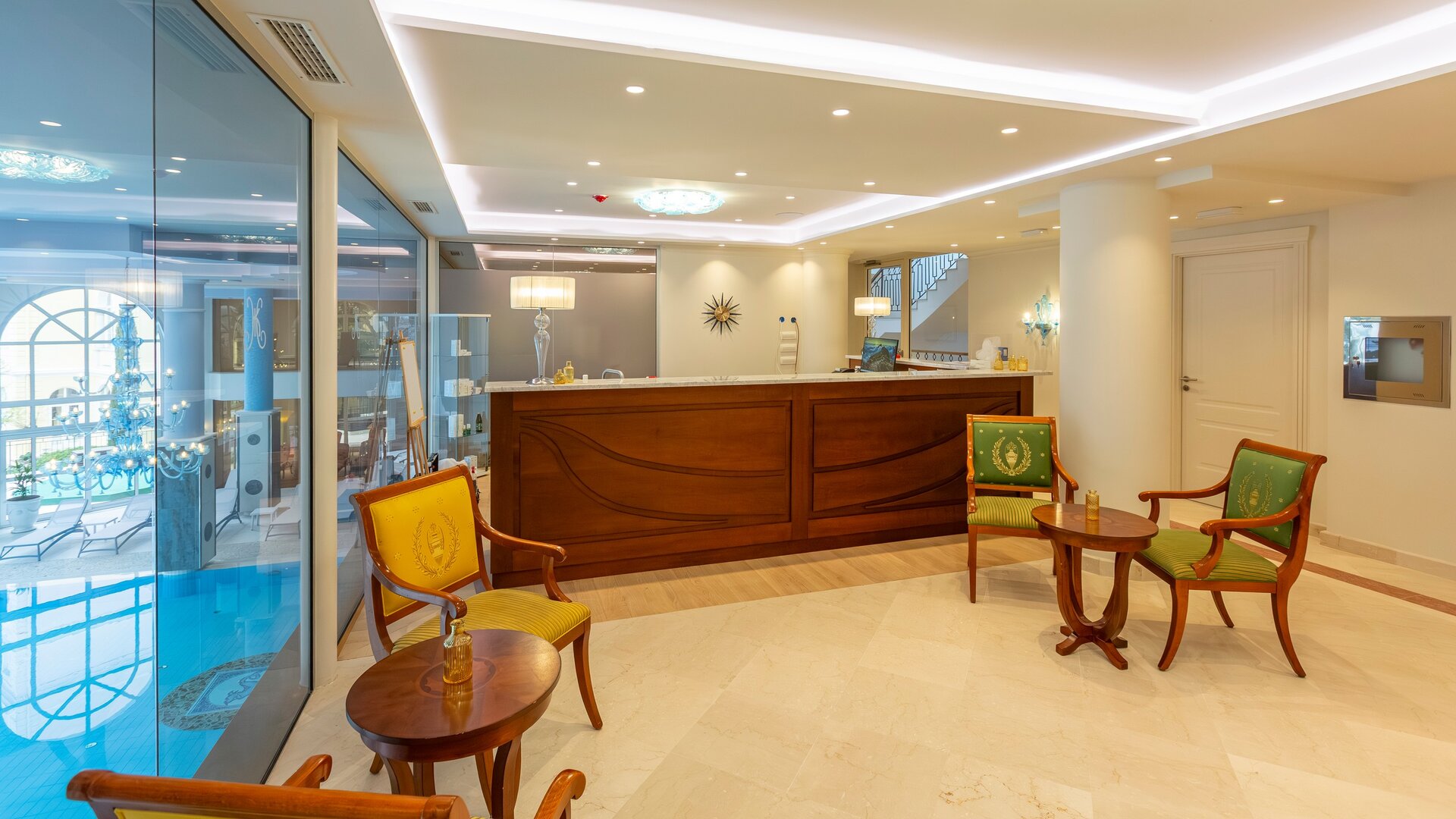 Helle Hotelrezeption mit einem eleganten Empfangstresen, umgeben von modernen Stühlen und Tischen. Große Fenster lassen Tageslicht herein und bieten einen Blick auf den Außenbereich. Die Atmosphäre ist einladend und freundlich.