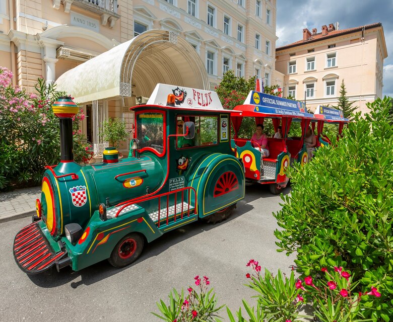 Bunte Touristenbahnen stehen vor einem eleganten Hotelgebäude, umgeben von üppigem Grün und blühenden Pflanzen. Der Himmel ist teilweise bewölkt, was eine einladende Atmosphäre schafft.
