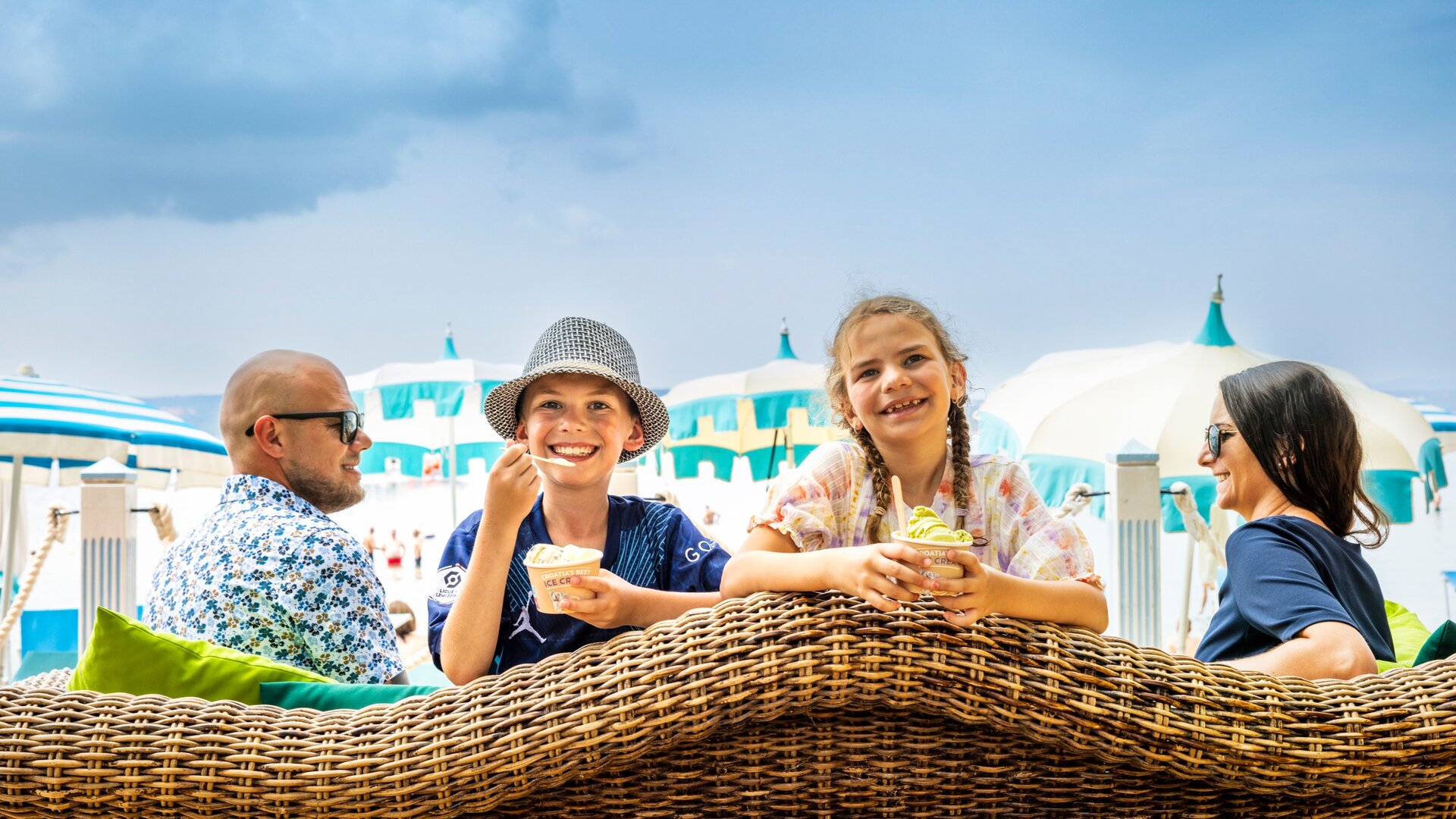 Zwei Kinder genießen Eiscreme auf einer gemütlichen Sitzgelegenheit am Strand, während im Hintergrund Sonnenschirme und eine entspannte Atmosphäre zu sehen sind. Ein Erwachsener sitzt ebenfalls in der Nähe, was die familiäre Stimmung unterstreicht.
