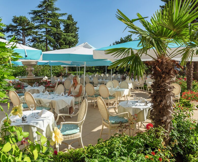 Ein elegantes Restaurant im Freien mit weißen Tischen und Stühlen, umgeben von üppigen Pflanzen und Palmen. Sonnenschirme bieten Schatten, während eine Frau entspannt an einem Tisch sitzt. Der Himmel ist klar und blau, was eine einladende Atmosphäre schafft.
