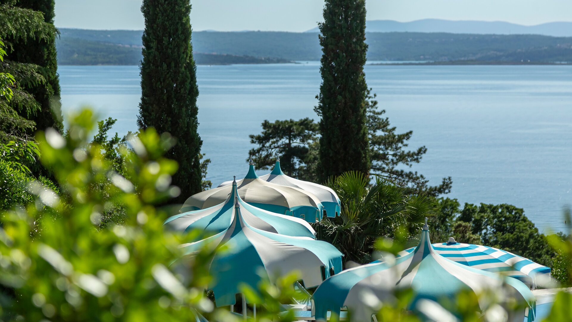 Blick auf den ruhigen See mit grünen Bäumen im Hintergrund und eleganten, gestreiften Sonnenschirmen im Vordergrund, die eine entspannte Atmosphäre schaffen.