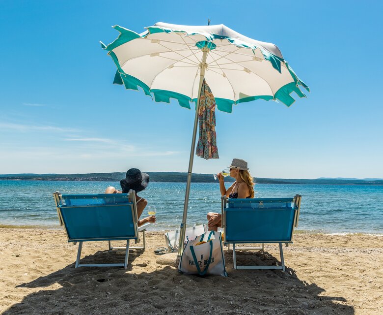Zwei Frauen sitzen unter einem großen Sonnenschirm am Strand. Sie entspannen auf blauen Liegen, während das klare Wasser sanft an den Strand plätschert. Der Himmel ist strahlend blau und die Atmosphäre ist entspannt und sonnig.