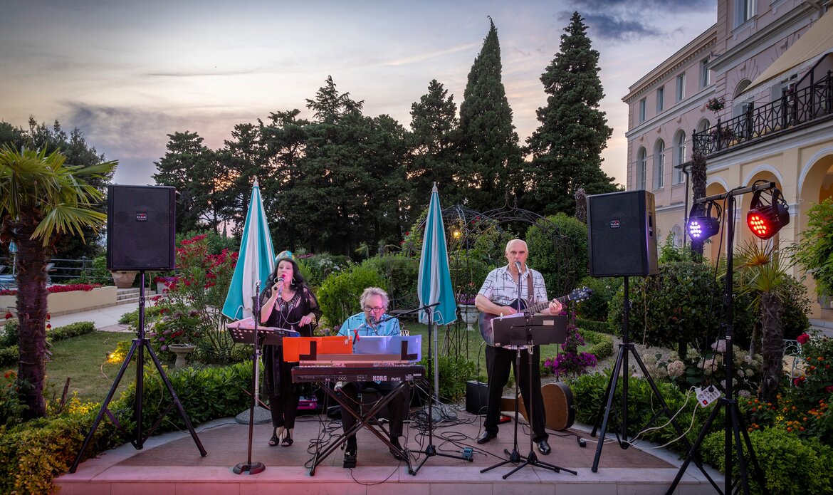 Eine Live-Band spielt im Freien auf einer Terrasse, umgeben von üppigem Grün und bunten Pflanzen. Die Musiker, darunter eine Sängerin und zwei Instrumentalisten, genießen den Abend mit sanftem Licht und einer entspannten Atmosphäre.