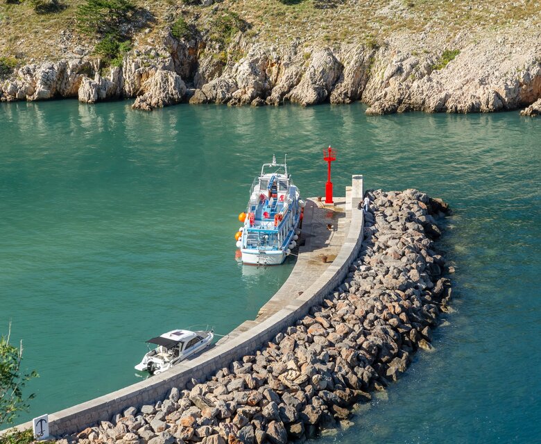Ein Boot liegt an einem kleinen Steg in einer ruhigen Bucht, umgeben von steilen Felsen. Ein rotes Leuchtfeuer steht am Ende des Stegs, während ein weiteres kleines Boot in der Nähe ankert. Das Wasser ist klar und grünlich-blau.