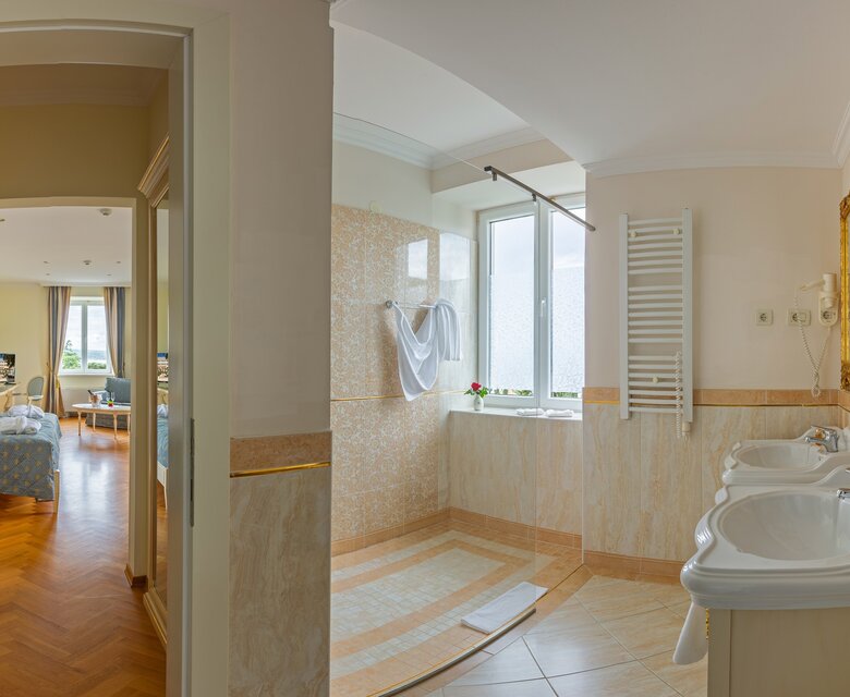 Ein helles Badezimmer mit zwei Waschbecken, einem großen Fenster und einem Handtuchwärmer. Im Hintergrund ist ein angrenzendes Schlafzimmer mit einem blauen Bett und großen Fenstern sichtbar. Der Raum hat einen eleganten, modernen Stil.
