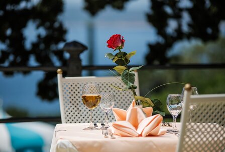 Ein elegant gedeckter Tisch im Freien mit einer roten Rose in der Mitte, umgeben von Gläsern und Besteck. Im Hintergrund ist ein schöner Ausblick auf das Wasser zu sehen, was eine romantische Atmos... Ein elegant gedeckter Tisch im Freien mit einer roten Rose in der Mitte, umgeben von Gläsern und Besteck. Im Hintergrund ist ein schöner Ausblick auf das Wasser zu sehen, was eine romantische Atmosphäre schafft.