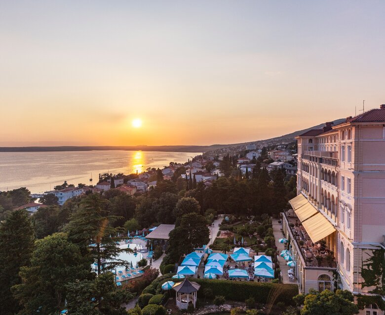 Blick auf ein Hotel am Meer bei Sonnenuntergang. Die Landschaft zeigt einen ruhigen Strand, grüne Bäume und einen Poolbereich mit blauen Sonnenschirmen. Die sanften Wellen des Wassers reflektieren das warme Licht der untergehenden Sonne.
