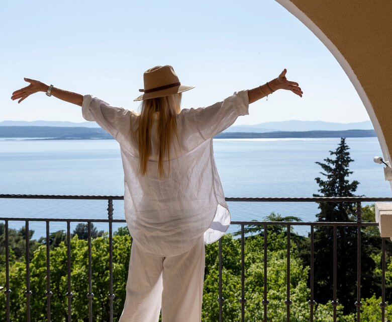 Eine Frau in einem weißen Outfit und einem Hut steht auf einem Balkon und breitet die Arme aus. Im Hintergrund erstreckt sich ein Blick auf das glitzernde Wasser und grüne Bäume, unter einem klaren blauen Himmel.