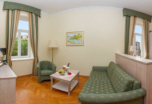 Helle Hotelzimmer mit einem gemütlichen Sofa, einem Sessel und einem Tisch. Große Fenster lassen viel Tageslicht herein. An der Wand hängt ein Landschaftsbild. Ein Fernseher steht auf einem Sideboa... Wunderschöne Zimmer im Familienhotel Crikvenica