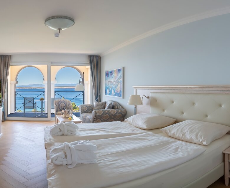 Ein helles Hotelzimmer mit einem großen Bett, einem gemütlichen Sessel und einem Schreibtisch. Große Fenster bieten einen Blick auf das Meer und den Balkon. Die Einrichtung ist modern und einladend, mit sanften Farben und natürlichem Licht.