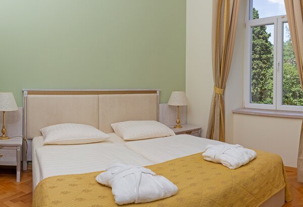 Helles, einladendes Hotelzimmer mit zwei Einzelbetten, einem gelben Überwurf und zwei Nachttischen mit Lampen. Ein großes Fenster mit Vorhängen lässt Tageslicht herein und bietet Blick auf die grün... Hotel mit Appartements für Familien, Kroatien - am Meer