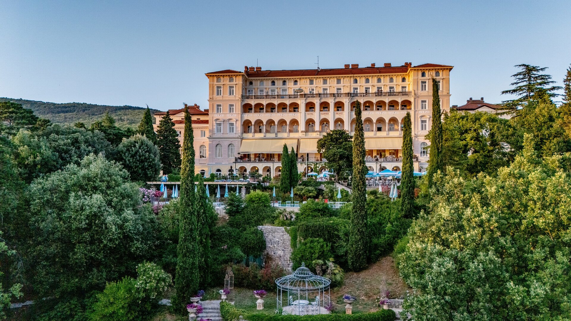 Ein elegantes Hotelgebäude mit mehreren Etagen, umgeben von üppigem Grün und gepflegten Gärten. Im Vordergrund sind Bäume und Blumenbeete zu sehen, während die Architektur des Hotels im Hintergrund hervorsticht. Die Atmosphäre ist ruhig und einladend.