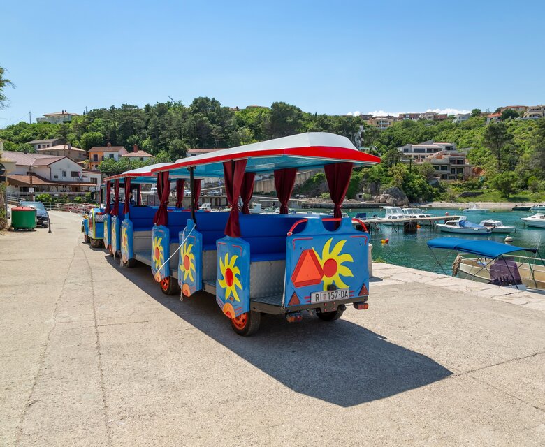 Bunte Touristenbahn auf einem gepflasterten Weg am Hafen, umgeben von Booten und grünen Hügeln. Im Hintergrund sind Wohnhäuser sichtbar, die in die Landschaft eingebettet sind. Der Himmel ist klar und sonnig.
