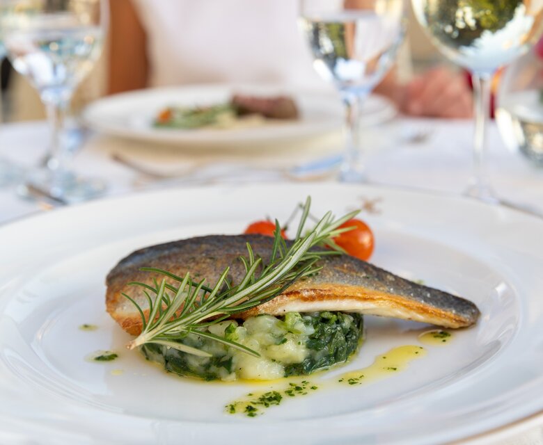 Ein elegant angerichteter Teller mit gebratenem Fisch, garniert mit Rosmarin und Kirschtomaten, auf einem Bett aus Kartoffel-Spinat-Püree. Im Hintergrund sind Gläser mit Wasser und ein weiterer Teller sichtbar, was eine gehobene Atmosphäre vermittelt.