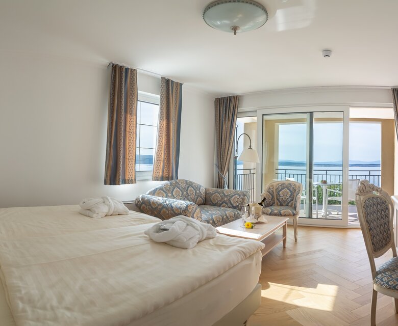 Helles Hotelzimmer mit Doppelbett, einem gemütlichen Sofa und einem Tisch. Große Fenster bieten einen Blick auf das Wasser. Ein Fernseher steht auf einem Schreibtisch, und der Raum ist modern und einladend gestaltet.