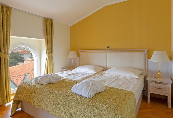Helles Hotelzimmer mit einem Doppelbett, weißen Bettdecken und einem goldenen Überwurf. An den Fenstern hängen gelbe Vorhänge. Neben dem Bett stehen zwei Nachttische mit Lampen. Durch das Fenster i... Appartements für Familienurlaub Crikvenica, Kroatien