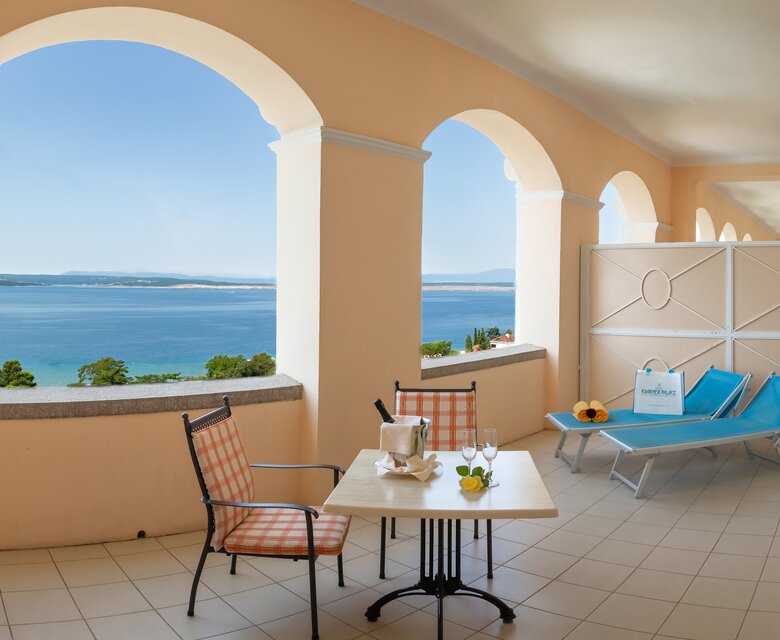 Ein Balkon mit Blick auf das Meer, ausgestattet mit einem kleinen Tisch, zwei Stühlen und Liegen. Die Wände sind in sanften Farben gehalten, und große Bögen lassen viel Licht herein. Die Atmosphäre ist entspannt und einladend.