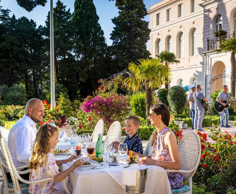 Eine Familie genießt ein Abendessen im Freien, umgeben von blühenden Pflanzen und Palmen. Im Hintergrund spielt eine Band, während die Sonne scheint und eine entspannte Atmosphäre schafft.