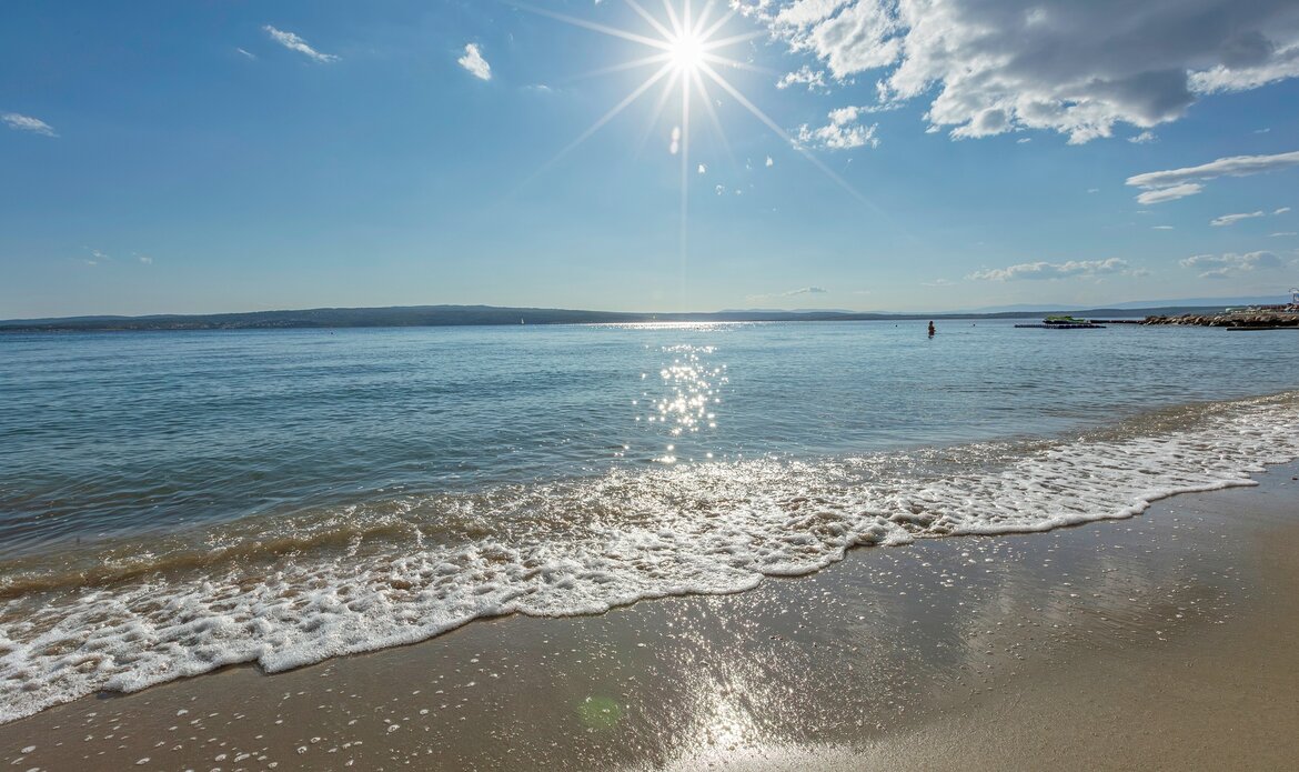 Ein sonniger Strand mit sanften Wellen, die an den Sand schlagen. Der Himmel ist klar mit einigen Wolken, und das Wasser glitzert im Sonnenlicht. Im Hintergrund ist eine ruhige Küstenlinie zu sehen.