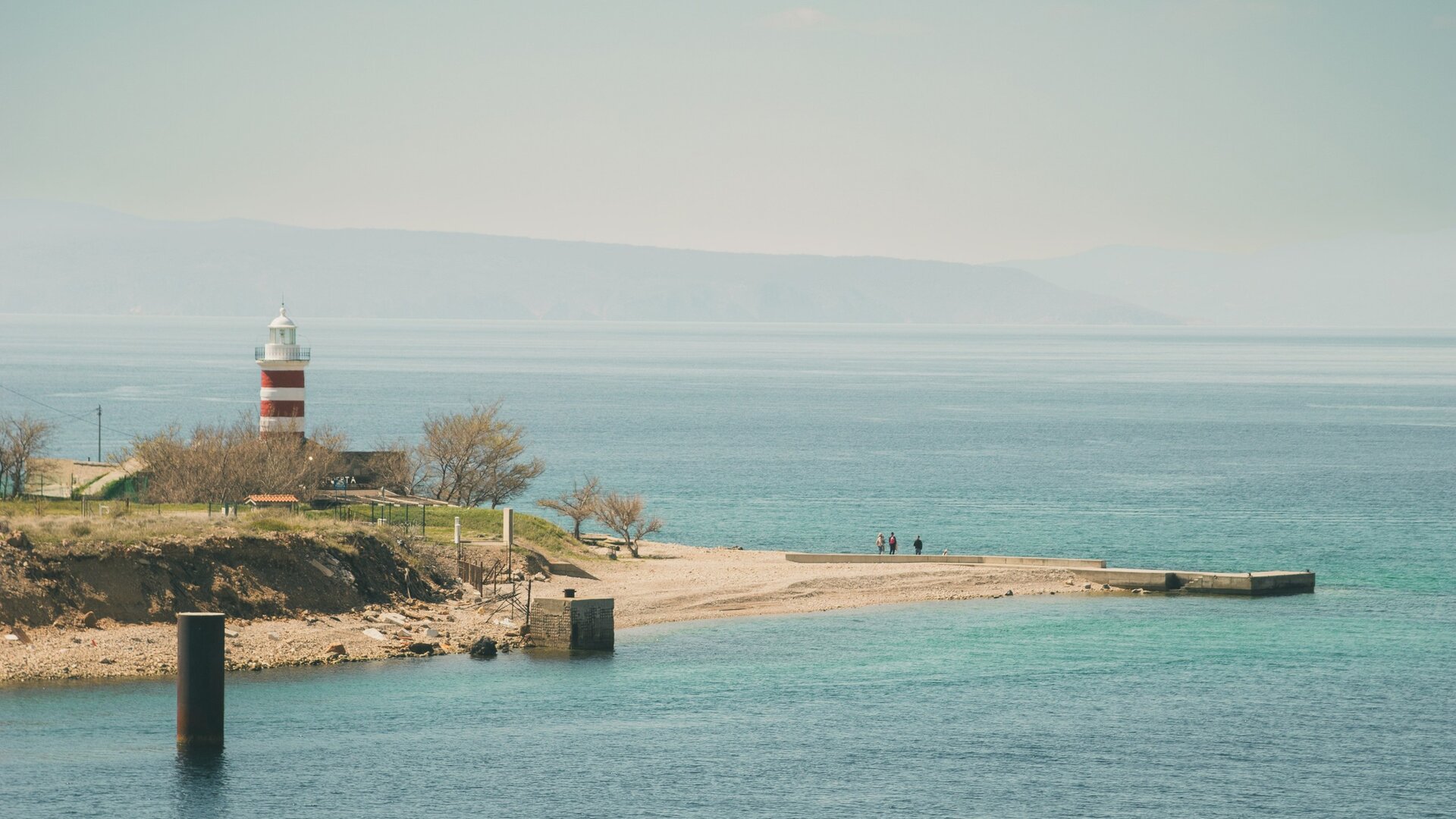 Ein malerischer Küstenblick mit einem rot-weißen Leuchtturm, der über das ruhige, türkisfarbene Wasser wacht. Im Hintergrund sind sanfte Hügel zu sehen, während einige Personen am Strand spazieren. Die Atmosphäre ist friedlich und einladend.