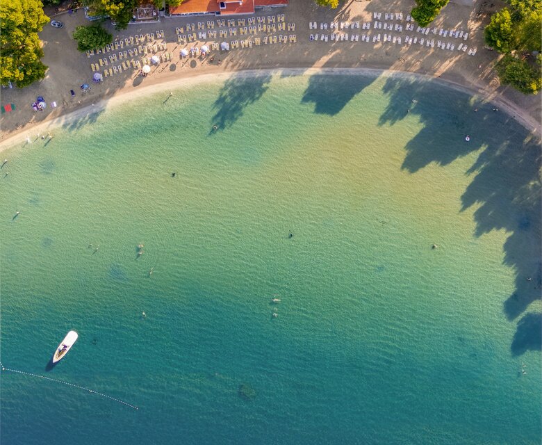 Luftaufnahme eines ruhigen Strandes mit klarem, türkisfarbenem Wasser. Am Ufer stehen Liegen, umgeben von Bäumen. Im Hintergrund sind einige Gebäude und ein Boot auf dem Wasser zu sehen. Die Atmosphäre ist entspannt und einladend.