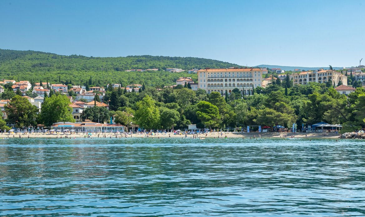Blick auf einen sonnigen Strand mit klarem Wasser, umgeben von grünen Bäumen und Hügeln. Im Hintergrund sind mehrere Gebäude und eine belebte Strandpromenade zu sehen. Die Atmosphäre ist entspannt und einladend.