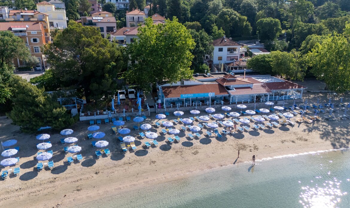 Ein Blick auf einen Strand mit vielen blauen Sonnenschirmen und Liegen. Im Hintergrund befindet sich ein Restaurant mit einer Terrasse, umgeben von Bäumen und Wohngebäuden. Das Wasser ist klar und ruhig, ideal für einen entspannten Tag am Meer.
