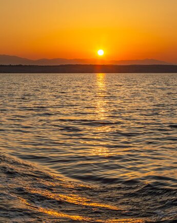 Sonnenuntergang über dem Meer mit ruhiger Wasseroberfläche und einer dunklen Küstenlinie am Horizont.
