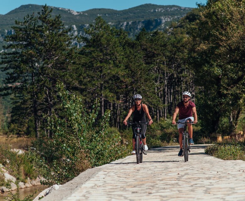 Zwei Radfahrerinnen fahren auf einem gepflasterten Weg entlang eines Flusses, umgeben von grünen Bäumen und Bergen im Hintergrund. Die Sonne scheint, und die Atmosphäre ist lebhaft und aktiv.